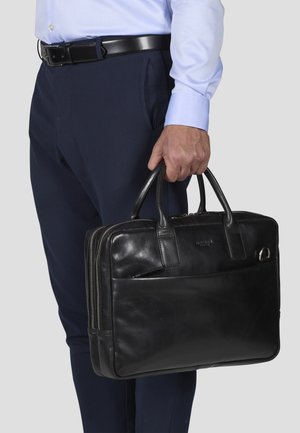 Schwarze Lederaktentasche mit zwei oberen Griffen, Reißverschluss und Fronttasche, die in einer Hand gehalten wird, kombiniert mit marineblauen Hosen.