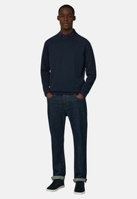 Jeune homme debout, les mains dans les poches, portant un pull marine, un jean bleu foncé avec des revers, et des baskets noires à semelles blanches.