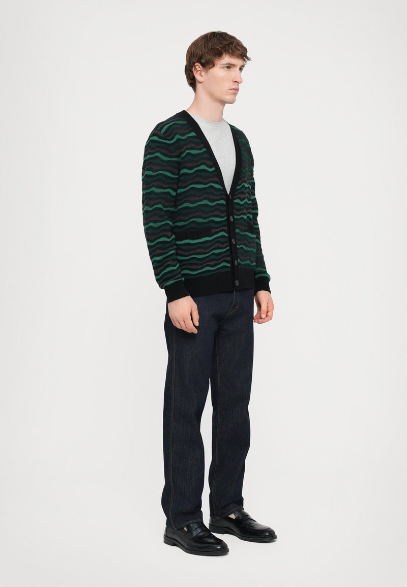 Black cardigan with green wave patterns, v-neckline, button closure, ribbed cuffs, worn over a gray t-shirt, paired with dark jeans and black shoes.