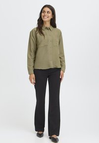 Donna in piedi e sorridente, che indossa una camicia verde oliva con bottoni, pantaloni neri a gamba larga e ballerine nere a punta, su uno sfondo bianco.