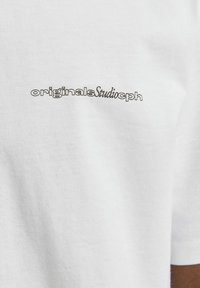 Balta medvilninė marškinėliai su juodu siuvinėtu logotipu "originalsStudio" kairėje pusėje. Minkšta tekstūra, trumpomis rankovėmis, paprastas dizainas.