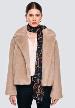 Beżowa kurtka z imitacji futra z szerokim kołnierzem, zestawiona z ciemnym szalikiem ozdobionym kolorowymi wzorami łańcuchów, noszona na jasnej, zapinanej na guziki koszuli.