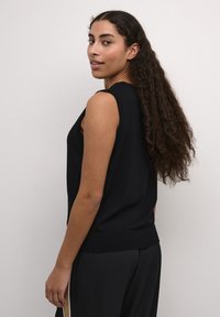 Zwarte mouwloze gebreide top met een ontspannen pasvorm, voorzien van een ronde hals en ribbels aan de onderkant. Model draagt het met zwarte broeken.