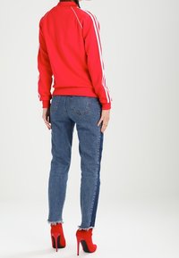 Červená sportovní bunda s bílými pruhy, doplněná modrými džínami s bočním pruhem a třepeným lemem, kombinovaná s červenými vysokými podpatky.