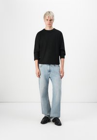 Pull en maille noir avec des manches trois-quarts, associé à un jeans évasé bleu clair et des chaussures en cuir noir. Fond uni.