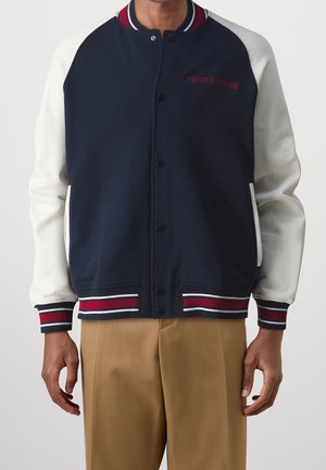 Hombre con chaqueta varsity azul marino y blanca con puños y cuello a rayas rojas, blancas y azul marino, combinada con pantalones marrón claro.