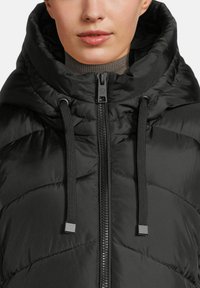 Woman wearing a black quilted puffer jacket with a high collar, front zipper, and black drawstrings over a gray turtleneck sweater.