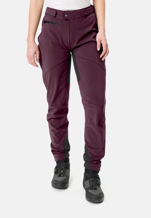 Frau trägt lila Outdoor-Hose mit schwarzen Patches und grau-schwarzen Wanderschuhen vor weißem Hintergrund.