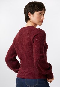 Pull en tricot bordeaux avec un motif texturé, des poignets et un ourlet à côtes. Le design comprend un col rond et des épaules légèrement bouffantes.