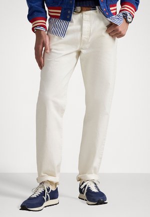 Weiße Denim-Jeans mit geradem Schnitt und feiner Textur. Kombiniert mit dunkelblauen Sneakers und einer blau-roten Collegejacke.