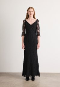 Robe en dentelle noire avec de longues manches transparentes, décolleté en V et corsage ajusté. Elle s'étend jusqu'au sol avec un ourlet festonné.