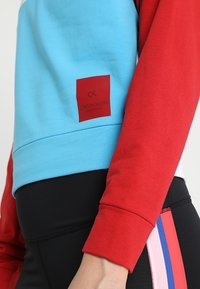 Calvin Klein sweatshirt featuring a sky blue body and red sleeves, made of soft fabric. Includes a red logo patch on the side.