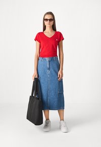 Red fitted t-shirt, blue denim midi skirt with pockets, black tote bag, and white sneakers. Accessories include dark sunglasses and hoop earrings.
