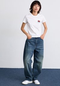 Camiseta de algodón blanca con un gráfico de cereza en el lado izquierdo del pecho, combinada con unos pantalones vaqueros de mezclilla azul holgados con una textura desgastada y zapatillas blancas.