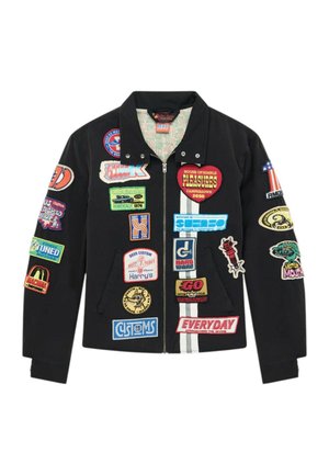 Hombre con una chaqueta negra cubierta de parches de colores sobre una camisa marrón de malla y vaqueros azul claro con rayas amarillas.