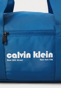 Blauer Duffle Bag aus glattem Stoff, mit einer Reißverschlusstasche und fettem weißen Text, der "calvin klein" und "West 39th Street" liest.