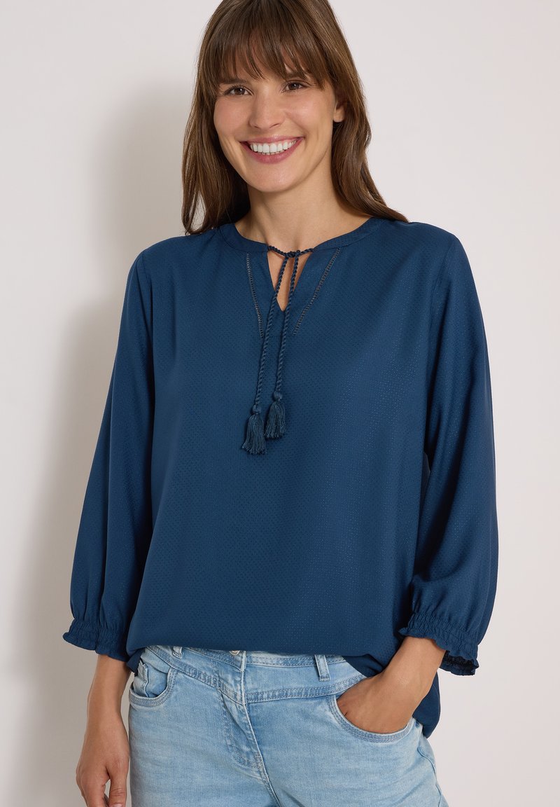Femme souriante aux cheveux bruns portant un chemisier bleu à manches longues avec des liens à pompons et un jean bleu clair, debout avec une main dans la poche.