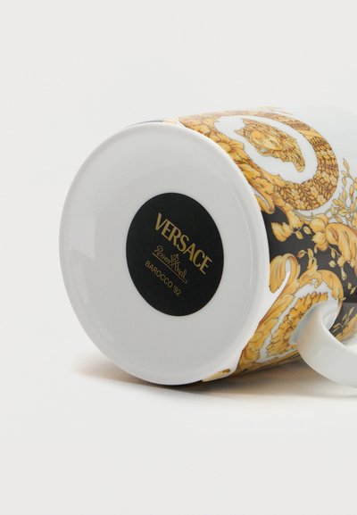 Λευκή κεραμική κούπα με χρυσό λουλουδάτο μοτίβο Barocco '92 και τη σήμανση Versace Rosenthal σε μαύρο κύκλο στη βάση, εμφανίζεται ξαπλωμένη στο πλάι.