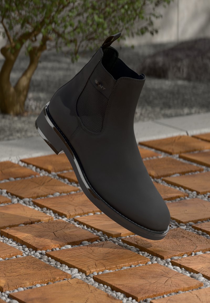 Bota chelsea de cuero negro con paneles laterales elásticos, acabado suave y suela brillante, situada sobre una superficie de piedra estampada.