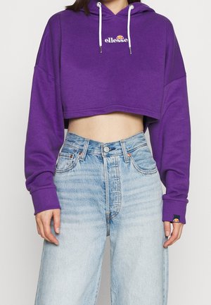 Person trägt ein lila kurzes Hoodie mit weißen Kordeln und hellblauen hoch taillierten Jeans vor einem einfarbigen Hintergrund.