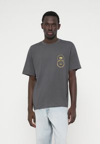 Hallide hallitus värvi puuvillane t-särk, ümmarguse kaelusega, lühikeste varrukatega ja vasakul rinnal kollane trükitud logo koos tähtedega.