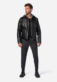 Schwarze Lederjacke mit grauem Kapuzenansatz, ausgestattet mit zwei Brusttaschen und Reißverschluss, kombiniert mit grauen Hosen und schwarzen Stiefeln.