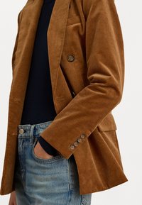 Bruine corduroy blazer met een dubbelzijdig ontwerp, voorzien van een notch-kraag en vier knopen op de manchetten, gecombineerd met blauwe denim jeans.