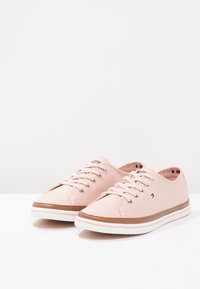Un paio di sneakers in tela rosa chiaro con suole bianche, rifiniture marroni e un piccolo logo rosso, bianco e blu sul lato e sul tallone interno.