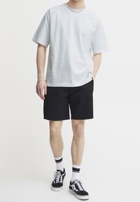 Homme portant un t-shirt blanc uni à manches courtes, un short noir, des chaussettes blanches avec des rayures noires et des baskets noires avec des lacets blancs.