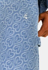 Tejido de mezclilla azul con un patrón de corazones en relieve, que cuenta con un logo bordado en blanco y una etiqueta lateral para ajuste o estilización.