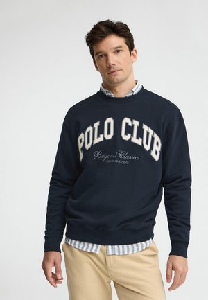 Homme portant un sweat-shirt bleu marine avec l'inscription "POLO CLUB", une chemise rayée bleu clair en dessous et un pantalon beige, debout devant un fond uni.
