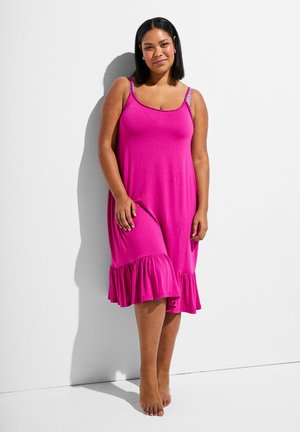 Keskmise nahatooniga naine, kellel on mustad õlgadeni juuksed, kannab varrukateta erkroosat keskmise pikkusega kleiti rippuva äärega, paljajal, seistes valge seina ääres.