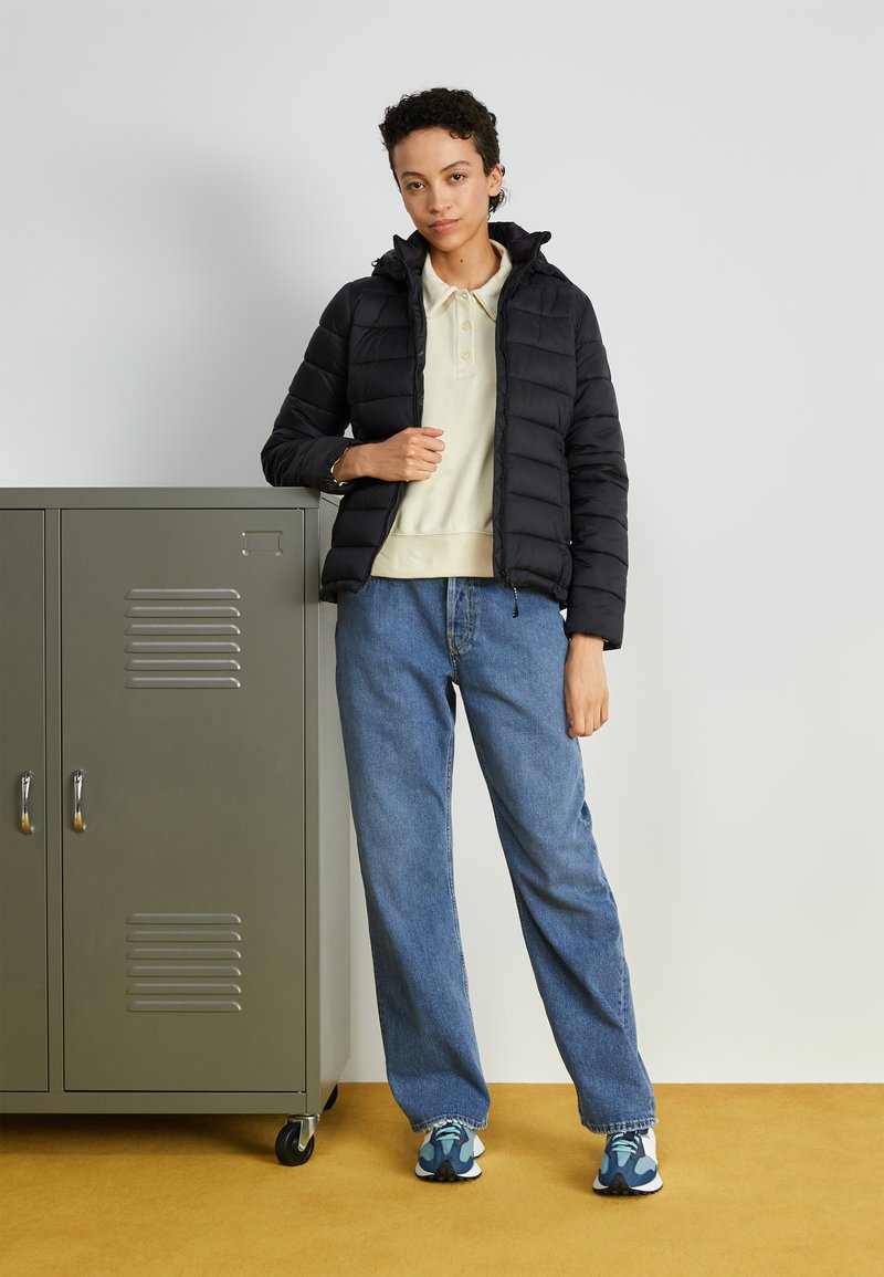 Chaqueta de plumón negra con capucha, camiseta polo crema, jeans azules holgados y zapatillas deportivas azules con estampado, ubicados al lado de una taquilla gris.