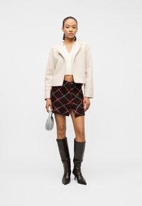 Γυναίκα με κοτσίδες, που φοράει κρεμ κοντό μπουφάν, λευκό πλεκτό τοπ, καρό φούστα κουμπωτή, μαύρες μπότες μέχρι το γόνατο και κρατά μικρή μπλε τσάντα.