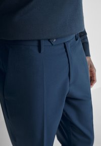 Muškarac u tamnoplavim krojenim hlačama i odgovarajućem tamnoplavom džemperu, prikazuje područje struka i gornjeg dijela nogu.