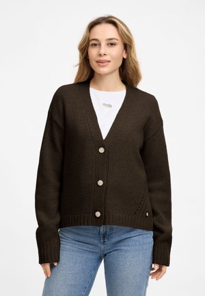 Frau mit blondem Haar, die einen dunkelbraunen, geknöpften Cardigan über einem weißen Hemd und blauen Jeans trägt, steht vor weißem Hintergrund.