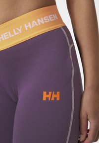 Paarse sportleggings met een gestructureerd patroon, voorzien van een brede oranje tailleband met de tekst "HELLY HANSEN" en een oranje logo op het been.