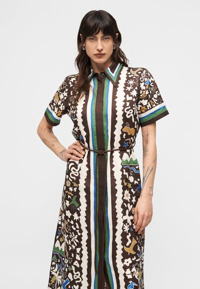 Γυναίκα με σκούρα μαλλιά που φοράει πολύχρωμο φόρεμα με κάθετες ρίγες σε πράσινο, μπλε και καφέ χρώμα και διάφορα αφηρημένα σχήματα, ποζάροντας με το ένα χέρι στη μέση.