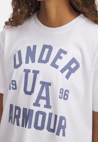 Camiseta de algodón blanca con gran texto en azul que dice "UNDER ARMOUR" junto a los números "1996" y "UA". Cuello redondo, corte relajado.