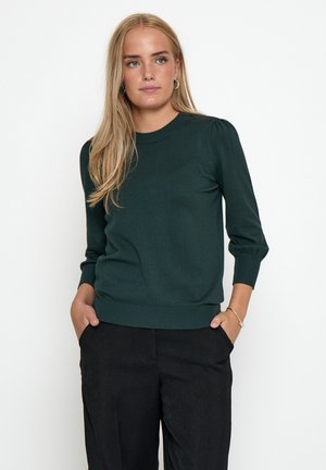 Vrouw met lang blond haar, gekleed in een donkergroen top met lange mouwen en een zwarte broek, met handen in haar zakken, staand voor een witte achtergrond.