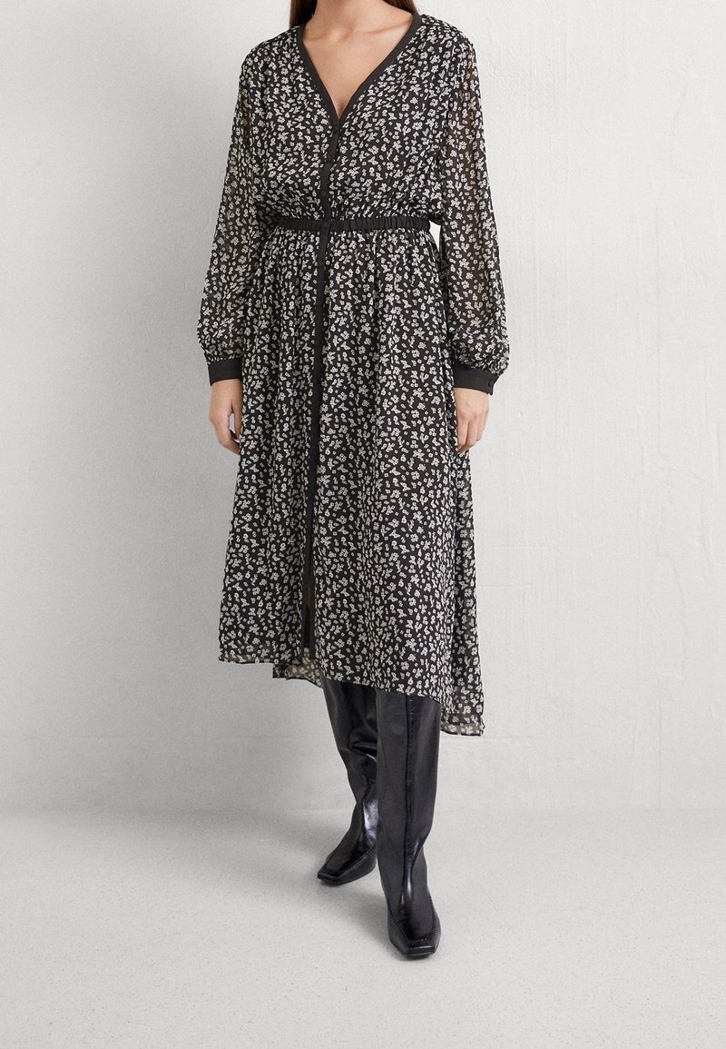 Femme portant une robe midi noire à motif floral blanc, col en V, manches longues transparentes, devant boutonné, et bottes noires montantes jusqu'au genou.