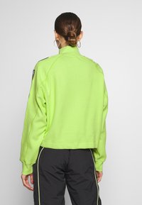 Felpa cropped verde neon con colletto alto, polsini a coste e dettagli neri sulle spalle. Tessuto liscio, vestibilità casual.