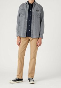 Camisa de mezclilla gris con botones a presión, dos bolsillos en el pecho, combinada con pantalones caqui y zapatillas negras con suelas y cordones blancos.