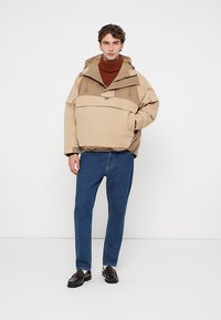 Veste pull beige avec une grande capuche et une poche avant, associée à un pantalon en denim bleu et des chaussures sombres. Le matériau semble texturé et durable.