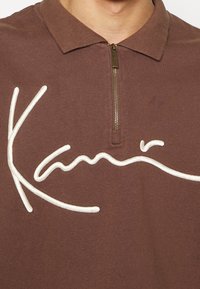 Polo marrone con colletto zip, caratterizzato da testo corsivo ricamato bianco "Kani" sulla parte anteriore. Tessuto morbido in cotone.