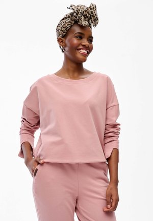 Uśmiechnięta kobieta ubrana w jasnoróżowy, długi rękaw, spodnie oraz z rękami w kieszeniach, z opaską w cętki i kolczykami kołami.