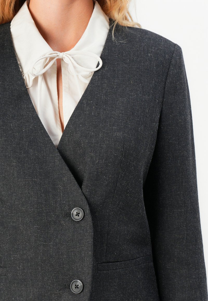 Blusa bianca con colletto a fiocco annodato sotto un blazer grigio scuro con bottoni, su una persona con capelli biondi, su uno sfondo bianco semplice.