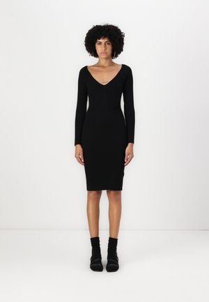 Black ribbed dress with a V-neckline and long sleeves, featuring a zipper detail at the chest. Worn with black ankle-length socks.