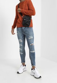 Pull en tricot orange, jean bleu déchiré, baskets blanches avec des accents noirs, et un sac bandoulière noir avec une fermeture éclair.