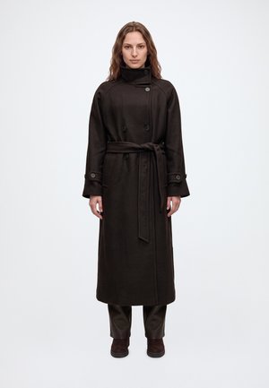 Une femme porte un long manteau brun foncé ceinturé à col montant, un pantalon foncé et des bottines, sur un fond clair et uni.